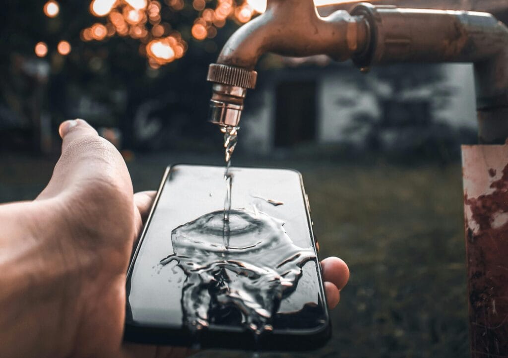 Unrecognizable male holding mobile phone under water pouring under tap in summer in evening time in countryside